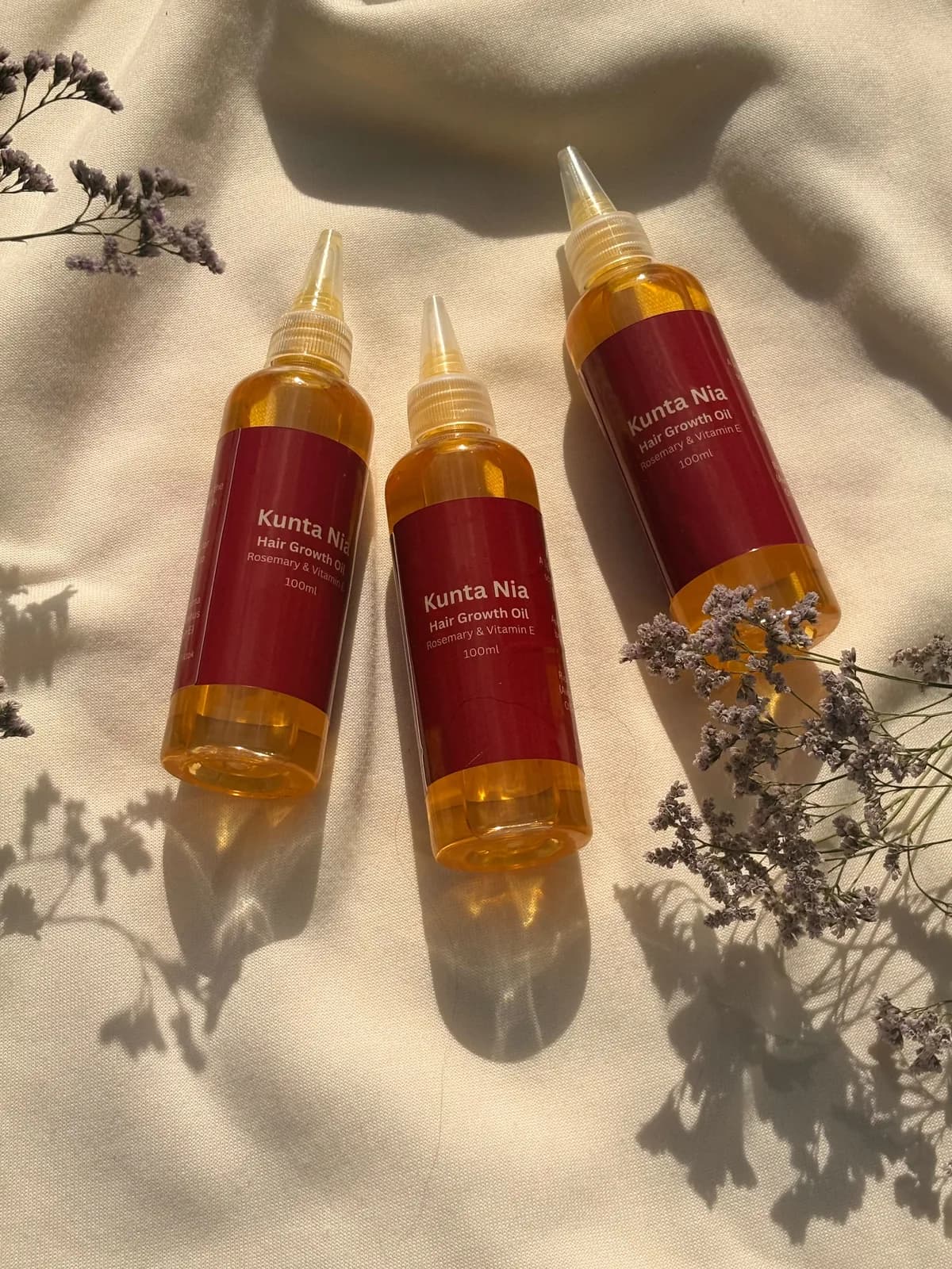 Three bottles of Kunta Nia Hair Growth Oil, Rosemary & Vitamin E, on cream cloth with dried flowers